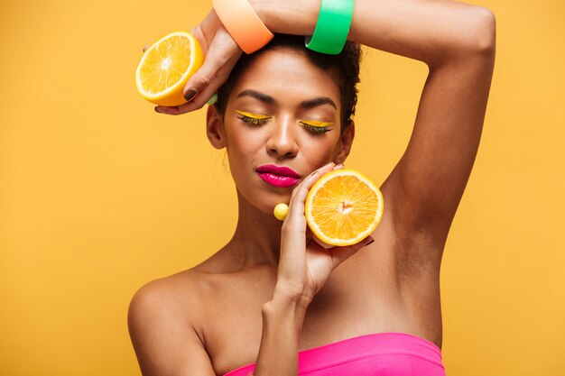 Retrato de mujer afroamericana sensual con los ojos cerrados sosteniendo dos partes de naranja en la cámara disfrutando de cítricos aislado, sobre pared amarilla