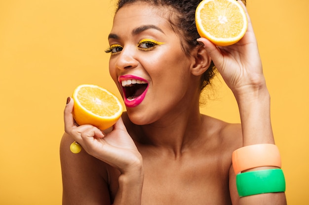 Retrato de mujer afroamericana sonriente con elegante maquillaje degustación jugosa naranja madura sosteniendo partes en ambas manos cerca de la cara aislada, sobre pared amarilla