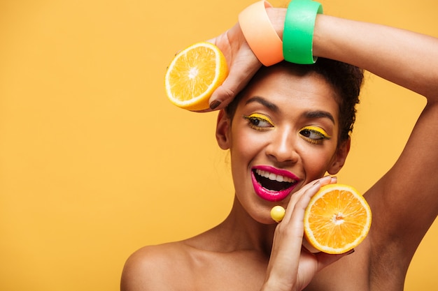 Retrato de mujer afroamericana sonriente con maquillaje elegante sosteniendo dos mitades de naranja jugosa en ambas manos aisladas, sobre pared amarilla