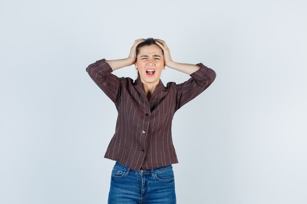 Retrato de mujer agarrando la cabeza con las manos mientras grita en camisa, jeans y mirando deprimido vista frontal