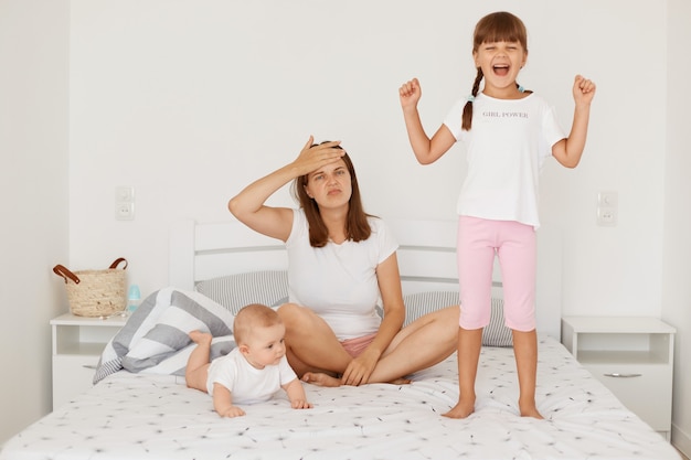 Retrato de mujer agotada con camiseta blanca sentada en la cama con sus dos hijas ruidosas, manteniendo la mano en la frente, cansada de pasar mucho tiempo con los niños.