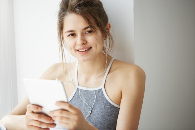 Retrato de la mujer alegre adolescente joven en auriculares que sonríe navegando navegando por Internet en la tableta que se sienta sobre la pared blanca.