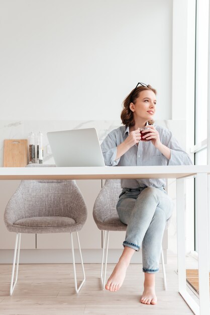 Retrato de mujer alegre en camisa a rayas y jeans sosteniendo una taza de té mientras está sentado en una silla suave en la cocina