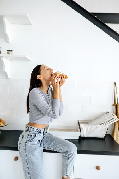 Retrato de mujer alegre come sabroso croissant en casa. Concepto de comida poco saludable.