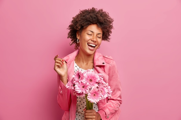 Retrato de mujer alegre y despreocupada sostiene ramo de flores de gerbera expresa emociones positivas cierra los ojos viste chaqueta de moda aislada en la pared rosa