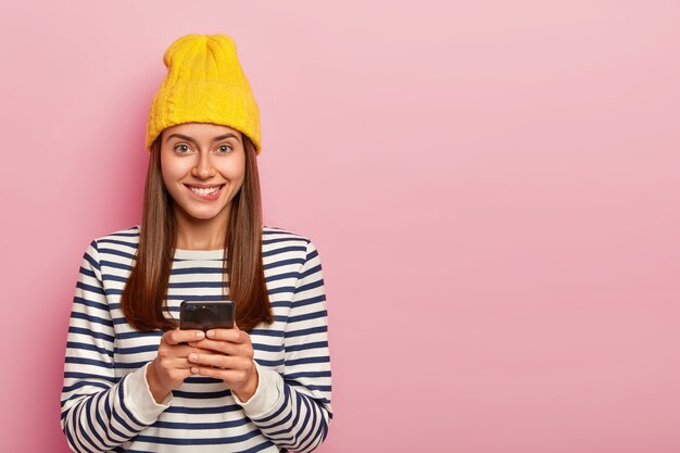 Retrato de mujer alegre encantada muerde los labios y mira felizmente a la cámara, usa un teléfono celular moderno, vestida con ropa elegante posa sobre un fondo rosado