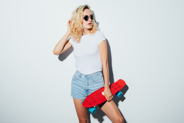 Retrato de una mujer alegre feliz en gafas de sol posando con patineta mientras está de pie sobre la pared blanca