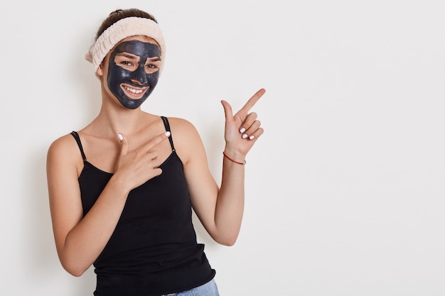 El retrato de una mujer alegre y feliz mejora la piel de su rostro, aplica una máscara exfoliante, está de buen humor, presenta modelos contra la pared blanca y señala con ambas manos a un lado.