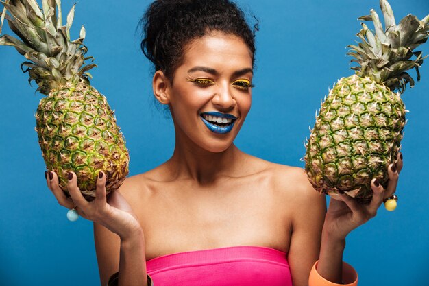 Retrato de mujer alegre mulata con maquillaje elegante guiñando un ojo mientras sostiene dos piñas jugosas en ambas manos aisladas, sobre pared azul