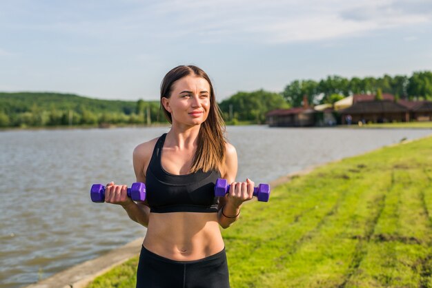 Retrato de mujer alegre en ropa deportiva haciendo ejercicio con mancuernas, al aire libre, con copyspace