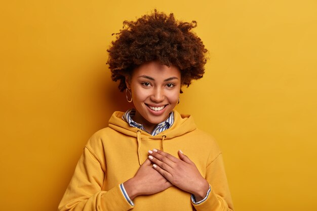 Retrato de mujer amigable de piel oscura hace gesto de gratitud, expresa agradecimiento por el cumplido recibido, usa sudadera con capucha, aislado sobre una pared amarilla, recibió sorpresa o elogio, siendo agradecido