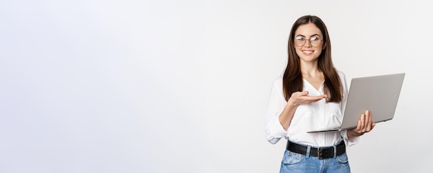 Foto gratuita retrato de mujer con anteojos sosteniendo una computadora portátil apuntando a la pantalla que muestra su trabajo en la computadora de pie