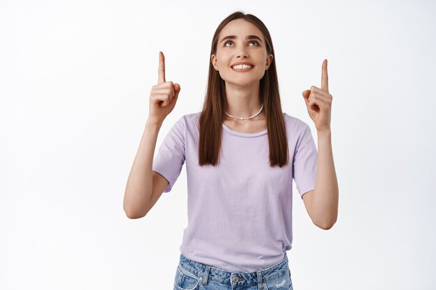 Retrato de mujer apuntando y mirando hacia arriba con sonrisa blanca satisfecha haciendo su elección, de pie sobre blanco en camiseta