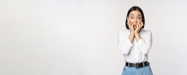 Retrato de una mujer asiática asustada viendo algo aterrador mordiéndose los dedos en las manos y mirando a la cámara asustada de pie sobre fondo blanco