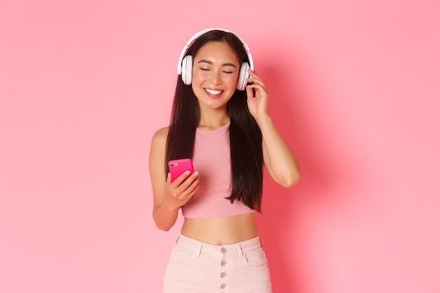 Retrato de mujer asiática atractiva despreocupada disfrutando de su canción favorita, cerrar los ojos para relajarse mientras escucha música en audífonos, sosteniendo el teléfono móvil y de pie sobre un fondo rosa.