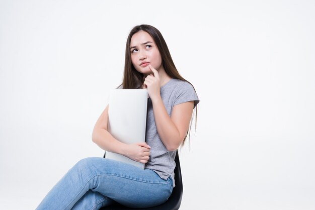 Retrato de una mujer asiática casual feliz sosteniendo la computadora portátil mientras está sentado en una silla sobre la pared blanca