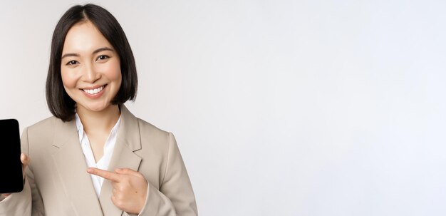 Retrato de mujer asiática corporativa que muestra la pantalla del teléfono móvil de la interfaz de la aplicación del teléfono inteligente de pie sobre fondo blanco