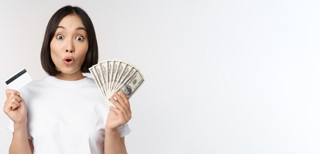 Retrato de mujer asiática con dinero en dólares y tarjeta de crédito que parece impresionada y asombrada de pie
