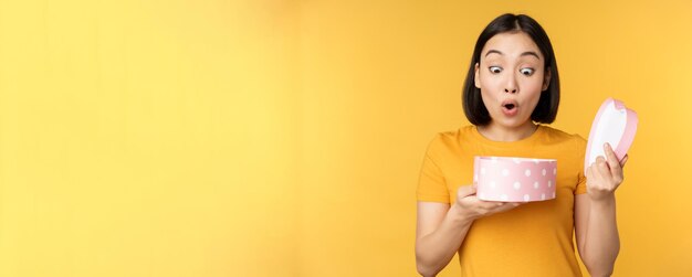 Retrato de mujer asiática emocionada caja de regalo abierta con cara feliz sorprendida de pie sobre fondo amarillo