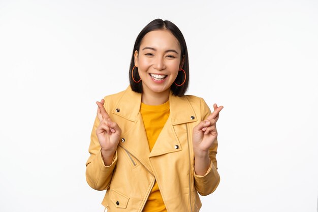 Retrato de una mujer asiática emocionada parece esperanzada deseando orar o rogar esperando noticias de pie sobre fondo blanco sonriendo entusiasta