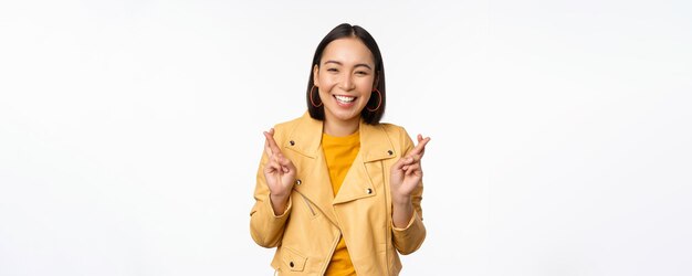 Retrato de una mujer asiática emocionada parece esperanzada deseando orar o rogar esperando noticias de pie sobre fondo blanco sonriendo entusiasta