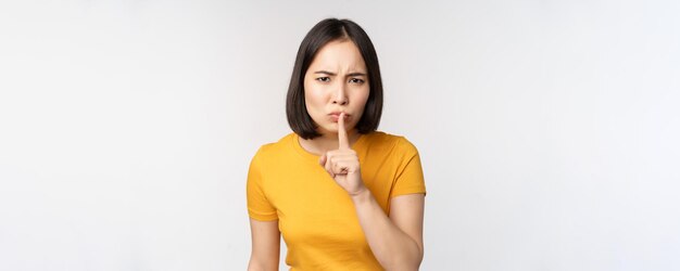 Retrato de una mujer asiática enojada que se calla presionando el dedo en los labios tabú deja de hablar sig mirando molesto regañando de pie sobre fondo blanco