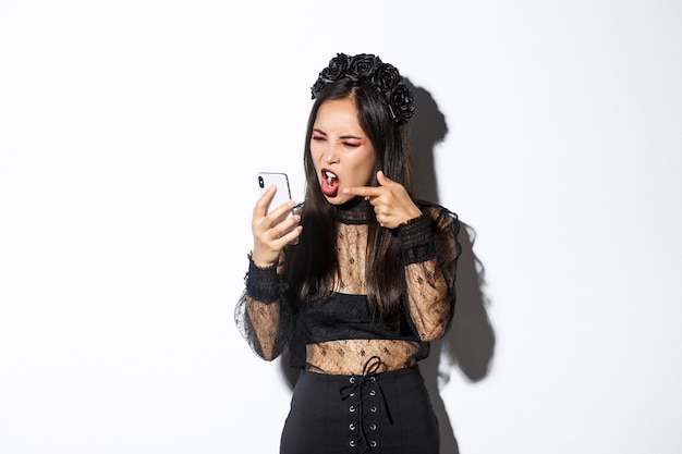 Retrato de mujer asiática enojada en traje de bruja discutiendo en la videollamada, gritando en el teléfono móvil y acusando a alguien, de pie sobre fondo blanco.