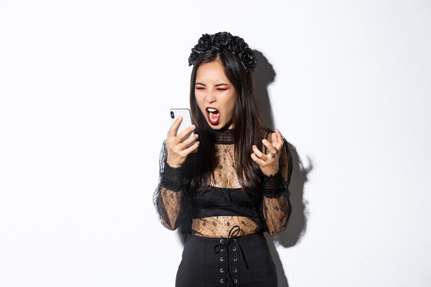 Retrato de mujer asiática enojada en traje de halloween que parece enojada, gritando en el teléfono móvil y haciendo muecas furiosas, de pie sobre fondo blanco.
