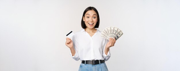 Retrato de mujer asiática entusiasta con dinero en efectivo y tarjeta de crédito sonriendo asombrada por el fondo blanco de la cámara