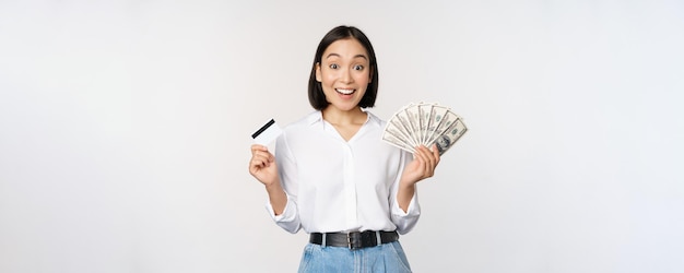 Retrato de mujer asiática entusiasta con dinero en efectivo y tarjeta de crédito sonriendo asombrada por el fondo blanco de la cámara