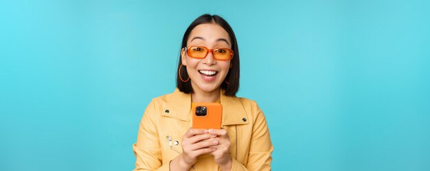 Retrato de una mujer asiática entusiasta con gafas de sol usando un teléfono móvil sonriendo y riéndose feliz sosteniendo un teléfono inteligente de pie sobre fondo azul