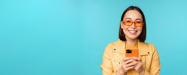 Retrato de una mujer asiática entusiasta con gafas de sol usando un teléfono móvil sonriendo y riéndose feliz sosteniendo un teléfono inteligente de pie sobre fondo azul