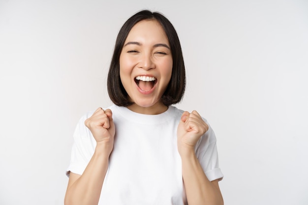 Retrato de mujer asiática entusiasta ganando celebrando y triunfando levantando las manos para alcanzar la meta o el éxito de pie sobre fondo blanco.