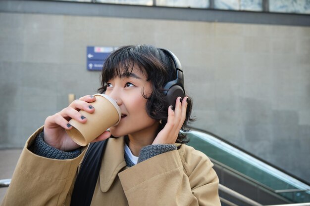 Retrato de mujer asiática con estilo en auriculares bebe café para llevar y sonríe disfruta de capuchino mientras