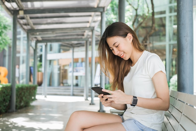 Retrato de la mujer asiática feliz atractiva que sostiene smartphone mientras que se sienta en el borde de la carretera en la ciudad