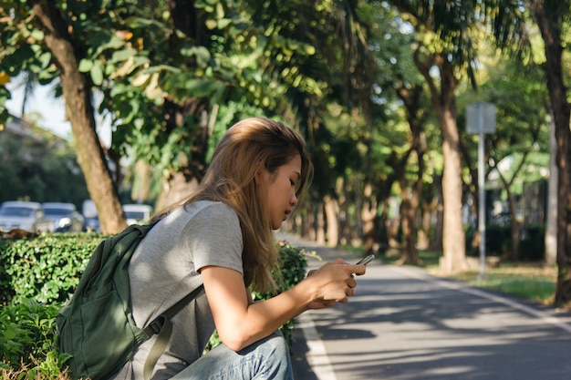 Foto gratuita retrato de la mujer asiática feliz atractiva que sostiene smartphone mientras que se sienta en el borde de la carretera en la ciudad