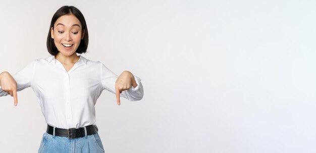 Retrato de una mujer asiática feliz señalando con el dedo hacia abajo y mirando debajo de un anuncio que muestra publicidad de pancartas de información de pie sobre fondo blanco