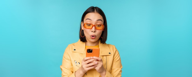 Retrato de una mujer asiática con gafas de sol mirando el teléfono móvil con una expresión de cara sorprendida de pie sobre fondo azul Copiar espacio