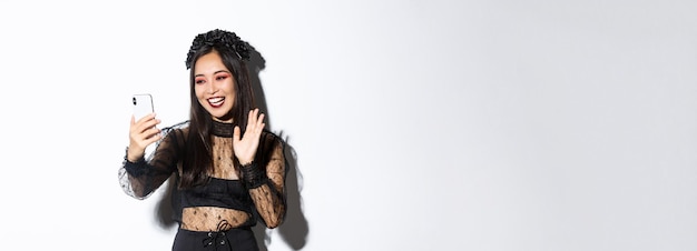 Foto gratuita retrato de mujer asiática hermosa y elegante en vestido de encaje gótico diciendo hola saludando con la mano en smartpho