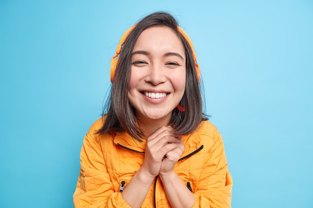 Retrato de mujer asiática hermosa feliz mantiene las manos juntas sonríe ampliamente disfruta de un buen sonido con auriculares modernos escucha música de la lista de reproducción lleva una chaqueta naranja aislada sobre una pared azul