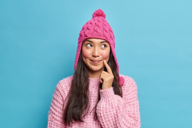 Retrato de mujer asiática hermosa pensativa vestida con gorro de invierno jersey de punto mira a un lado pensativamente modelos sobre pared azul