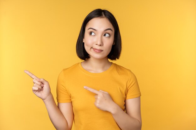 Retrato de mujer asiática intrigada mirando y señalando con el dedo a la izquierda en un anuncio que muestra algo interesante de pie sobre fondo amarillo
