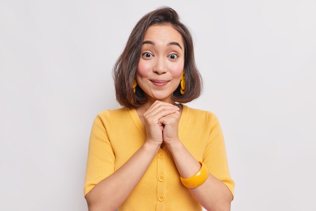 Retrato de mujer asiática joven de buen aspecto con piel sana de cabello natural dak mantiene las manos juntas debajo de la barbilla lleva un puente de aretes amarillos y una pulsera en el brazo aislado sobre una pared blanca