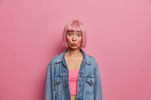 Retrato de mujer asiática joven con cabello bob rosa de moda, ha sorprendido expresión preocupada, escucha algo emocionante, vestida con una chaqueta de mezclilla de gran tamaño, posa en el interior. Chica oriental de moda