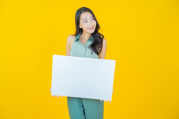 Retrato de mujer asiática joven hermosa con cartelera blanca vacía en la pared amarilla