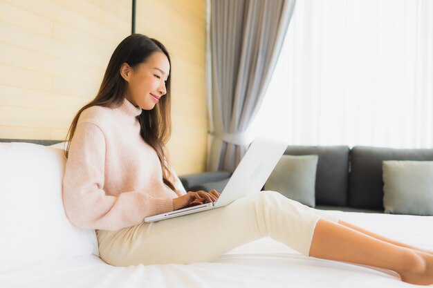 Retrato mujer asiática joven hermosa que usa la computadora portátil con el teléfono móvil en cama