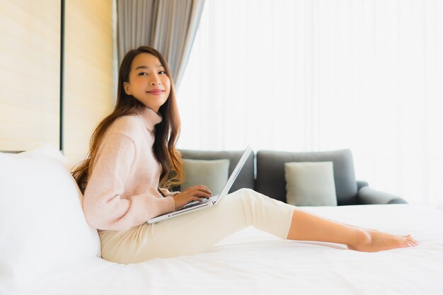 Retrato mujer asiática joven hermosa que usa la computadora portátil con el teléfono móvil en cama