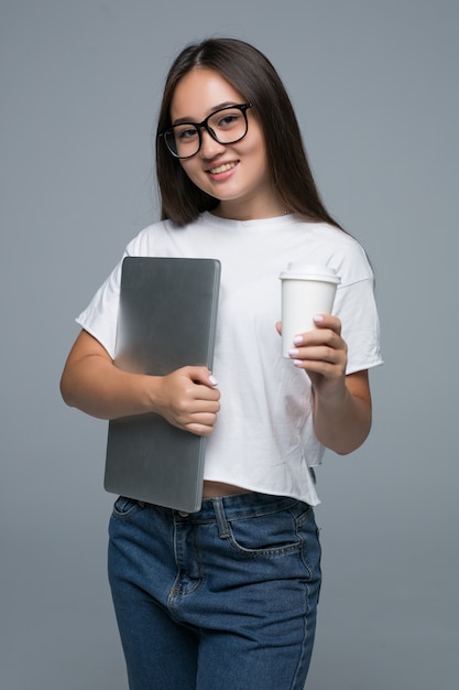 Foto gratuita retrato de una mujer asiática joven satisfecha sosteniendo una taza de café y una computadora portátil mientras camina y mira a la cámara sobre fondo gris