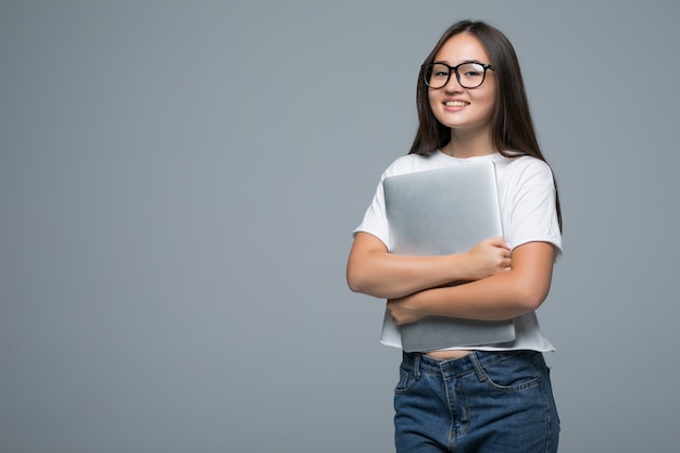 Retrato de una mujer asiática joven satisfecha sosteniendo una taza de café y una computadora portátil mientras camina y mira a la cámara sobre fondo gris