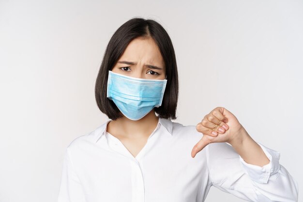 Retrato de una mujer asiática con mascarilla médica mostrando los pulgares hacia abajo con expresión de cara cansada decepcionada de pie sobre fondo blanco